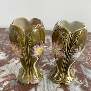 May include: Pair of green ceramic vases with floral designs. The vases have a bulbous shape with a flared opening and a slender base. The floral designs are in shades of pink, purple, and yellow.