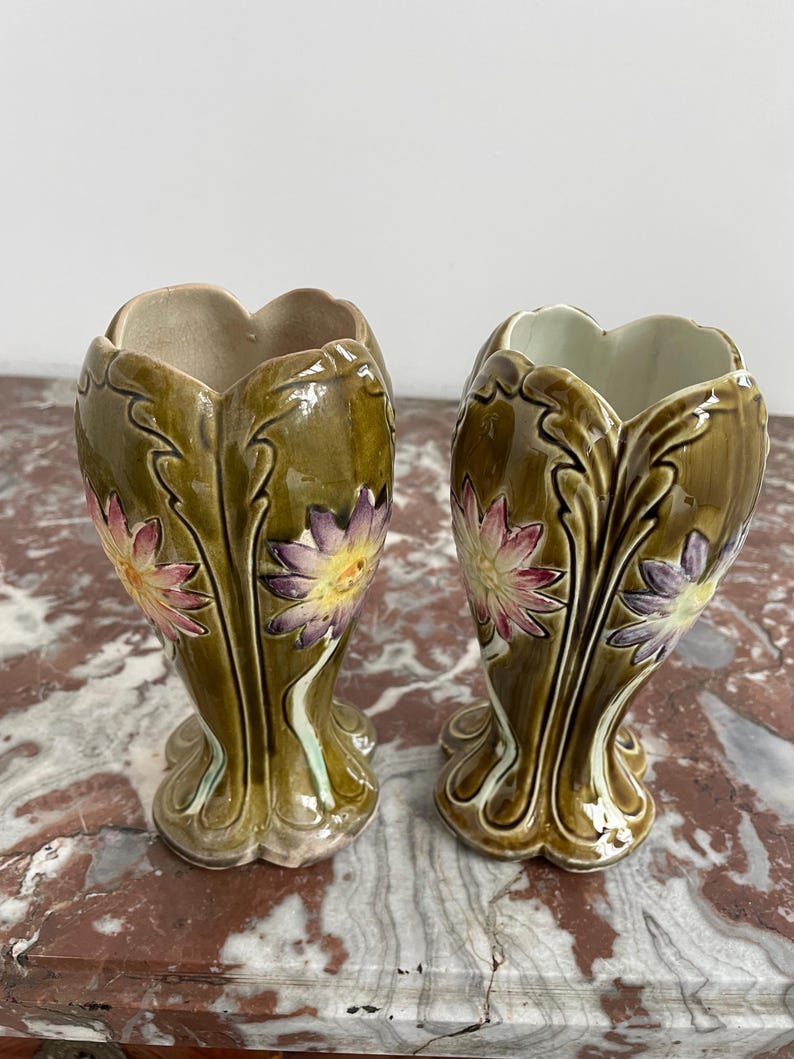May include: Pair of green ceramic vases with floral designs. The vases have a bulbous shape with a flared opening and a slender base. The floral designs are in shades of pink, purple, and yellow.
