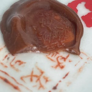 May include: A brown wax seal with embossed characters, resting on a white surface with red ink markings. The seal has a textured appearance and a slightly irregular shape. The image suggests a vintage or antique theme.