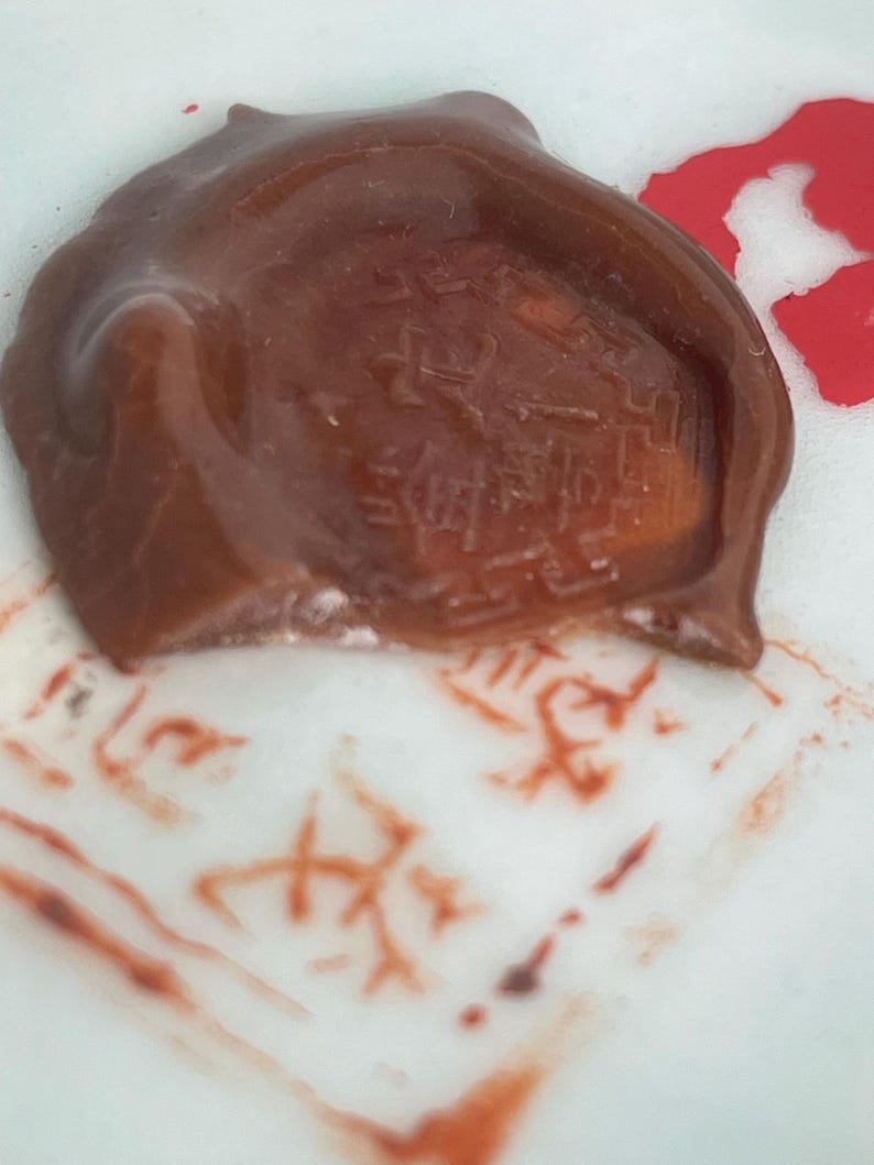 May include: A brown wax seal with embossed characters, resting on a white surface with red ink markings. The seal has a textured appearance and a slightly irregular shape. The image suggests a vintage or antique theme.