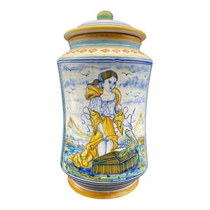 Op de afbeelding: Een decoratieve keramische pot met deksel, met een handgeschilderd ontwerp. De pot toont een vrouw in een gele jurk, een landschap en een vogel. Het kleurenpalet omvat blauw, geel en wit, met een gouden basis en een groene knop op het deksel.