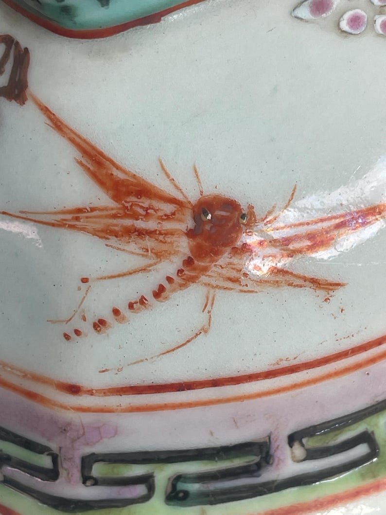 May include: Close-up of a white ceramic surface with a detailed insect illustration in shades of orange and red. The insect has delicate wings and a segmented body. The design is part of a larger decorative piece.
