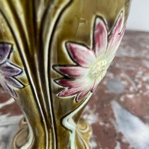 May include: A ceramic vase with a green and brown glaze. The vase is decorated with raised flowers in shades of pink, white, and purple. The vase has a flared base and a narrow neck.