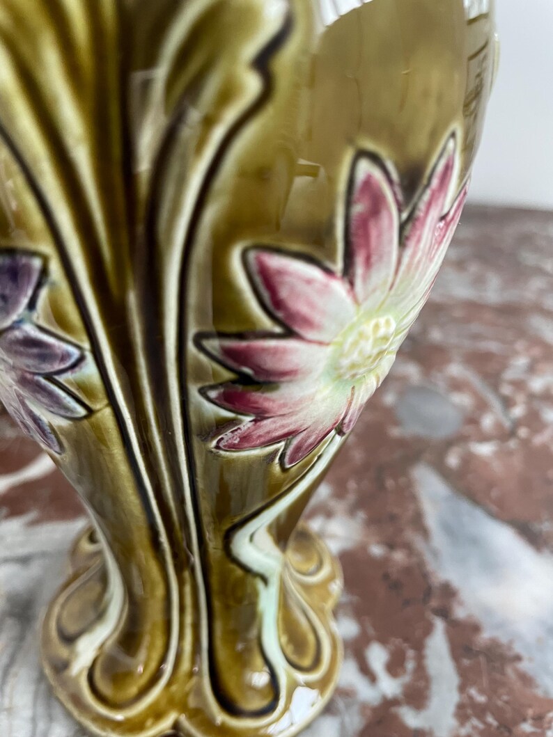 May include: A ceramic vase with a green and brown glaze. The vase is decorated with raised flowers in shades of pink, white, and purple. The vase has a flared base and a narrow neck.