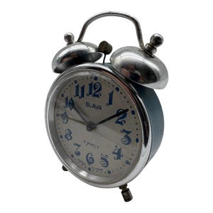 May include: A vintage Slava alarm clock with a metallic silver finish, featuring a round face with blue stylized numerals, black hands, and the text '11 JEWELS' and 'MADE IN USSR' printed on the dial, topped with two silver bells and a handle.