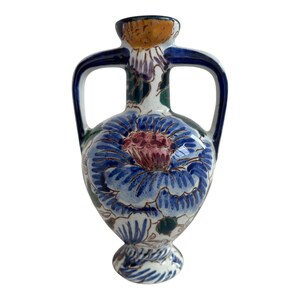 Può includere: Un piccolo vaso in ceramica bianca con disegni floreali blu e verdi. Il vaso ha due manici e un corpo arrotondato.