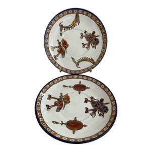 Op de afbeelding: Twee decoratieve keramische borden met een witte achtergrond, een blauwe rand en ingewikkelde ontwerpen in bruin, goud en blauw. De borden tonen een centraal motief van gestileerde figuren en sierlijke patronen.