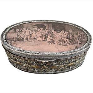 May include: An oval silver and gold box with an engraved image of a group of people gathered around a table. The box has a detailed floral design around the bottom edge.