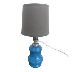 Puede incluir: Una pequeña lámpara de mesa de cerámica azul con una pantalla de tela gris.
