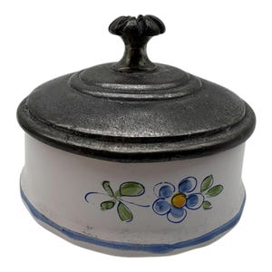 Può includere: Un piccolo contenitore rotondo in ceramica con coperchio in metallo. Il corpo bianco è decorato con fiori blu e foglie verdi, e una striscia blu accentua la base. Il coperchio ha una finitura scura e strutturata e una maniglia decorativa.