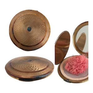 May include: A vintage gold-toned compact with a detailed zigzag pattern on the lid. The compact is round and has a small blue accent in the center. The interior reveals a mirror and a pink powder puff.