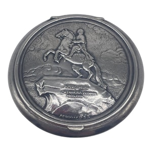 May include: A round silver-toned metal compact case featuring a raised relief design of a person riding a rearing horse on a rocky cliff. The text PETRO PRIMO CATHARINA SECUNDA MDCCLXXXII and the Cyrillic text ЛЕНИНГРАД are embossed on the surface.