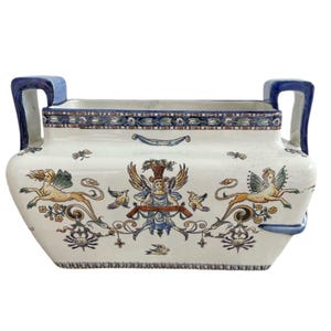 Puede incluir: Un macetero de cerámica blanca con detalles pintados en azul y dorado. El macetero presenta dos criaturas aladas con acentos florales y un diseño central de una figura con una corona.