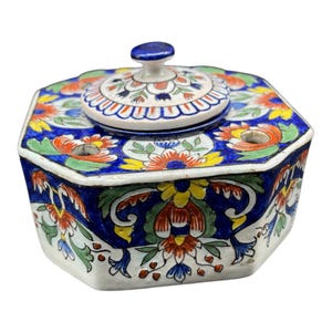 Könnte beinhalten: Ein handbemalter, achteckiger Keramikbehälter mit Deckel. Der Behälter ist mit einem Blumenmuster in Blau, Rot, Gelb und Grün verziert. Der Deckel hat einen kleinen blauen Knopf. Der Behälter dient wahrscheinlich zur Aufbewahrung oder Präsentation.