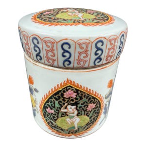 Può includere: Un vaso di ceramica bianco con coperchio, caratterizzato da intricati disegni dipinti a mano. Il corpo del vaso presenta un motivo centrale di un bambino su una foglia di loto, circondato da motivi floreali e geometrici dai colori vivaci. Il coperchio rispecchia il design.