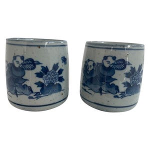 Könnte beinhalten: Zwei Keramikbecher mit blau-weißem Blumen- und Figuren-Design. Die Becher haben eine zylindrische Form und einen gesprenkelten, cremefarbenen Hintergrund. Das blaue Design zeigt Figuren, Blumen und Blätter.