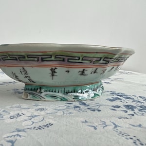 May include: A decorative, antique ceramic bowl with a scalloped rim. The bowl features a light green base with a decorative border of black, red, and purple geometric patterns. Chinese characters are visible on the side.