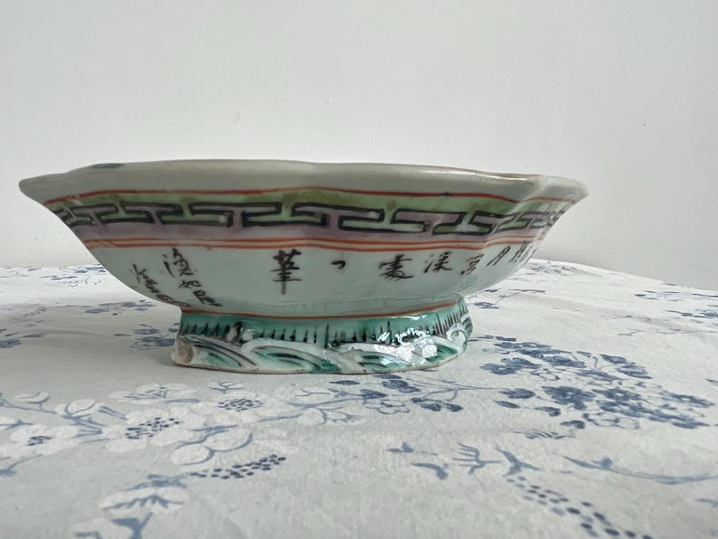 May include: A decorative, antique ceramic bowl with a scalloped rim. The bowl features a light green base with a decorative border of black, red, and purple geometric patterns. Chinese characters are visible on the side.