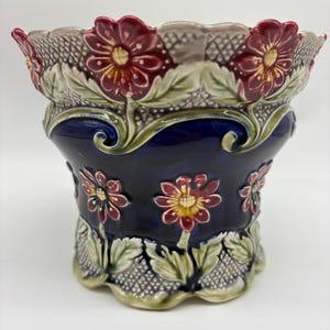 Peut inclure: Un cache-pot en céramique avec un corps émaillé bleu profond orné de fleurs rouges en relief avec des centres jaunes et des feuilles vertes. Les bords supérieur et inférieur présentent un rebord festonné et un motif quadrillé texturé.