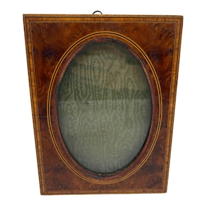 May include: A rectangular wooden picture frame featuring a rich brown burl wood veneer with a prominent oval center opening. The frame is accented by thin, dark inlaid border lines and contains a textured, muted green fabric backing material.