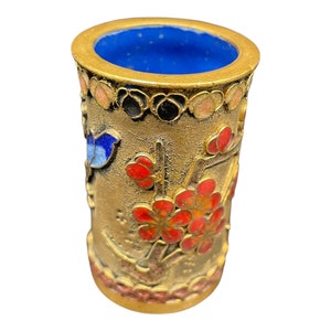 Puede incluir: Un pequeño jarrón cilíndrico de color dorado con un interior azul y diseños florales intrincados en esmalte rojo, naranja y azul. El jarrón está decorado con un patrón floral y un pequeño pájaro.