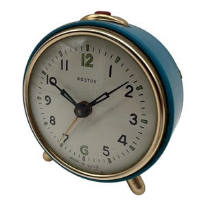 May include: A vintage teal alarm clock with a gold-colored bezel and legs. The clock face is off-white with black numerals and hands. The word "ROSTOV" is printed above the center. The clock reads 10:08.