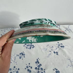 May include: A hand holds a vintage ceramic bowl. The bowl features a green interior and a decorative exterior with red, black, and green accents. The rim has a detailed pattern. The bowl is likely antique.