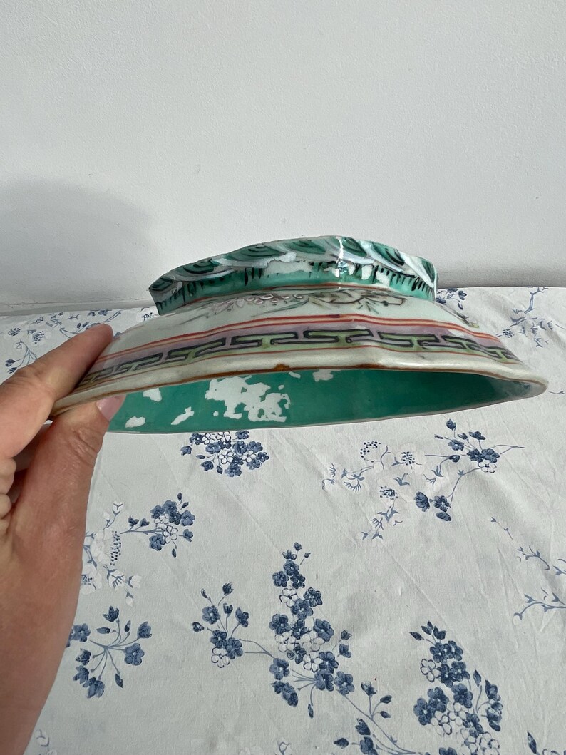 May include: A hand holds a vintage ceramic bowl. The bowl features a green interior and a decorative exterior with red, black, and green accents. The rim has a detailed pattern. The bowl is likely antique.