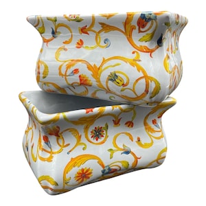 Könnte beinhalten: Zwei rechteckige Keramikbehälter in Weiß mit gewölbter Oberseite. Die Behälter sind mit einem sich wiederholenden Muster aus goldenen, orangefarbenen und blauen floralen Designs verziert. Die Behälter sind übereinander gestapelt.