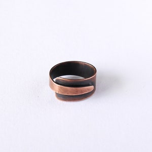 Può includere: Un anello in rame fatto a mano con un design aperto unico. L'anello presenta una fascia scura e ossidata con un accento in rame più chiaro a contrasto. Il design dell'anello è moderno e minimalista, adatto all'uso quotidiano.