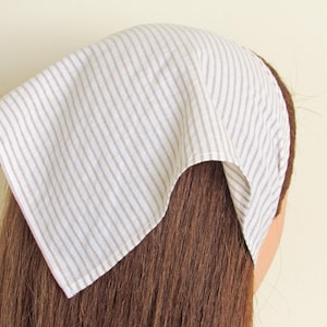 May include: A white and beige striped headscarf, folded and tied, covering the top of the head. The fabric has vertical stripes. The headscarf is made of a soft, lightweight material.