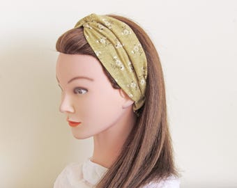 May include: A knotted headband in a light olive green fabric with a delicate white floral pattern. The headband is worn on a mannequin with brown hair and a white top. The fabric appears soft and likely made of cotton or a similar material.