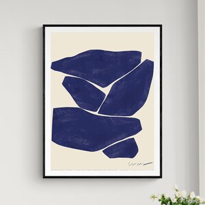 May include: Abstract art print featuring a composition of dark blue shapes on a cream background, framed in black. The artwork is signed in the lower right corner. The print is displayed in a room with a blue chair, white table, and a striped rug.