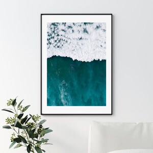 May include: A framed print featuring an aerial view of a turquoise ocean with a breaking wave. The white foam of the wave contrasts with the deep blue water. A surfer is visible in the water. The frame is black.