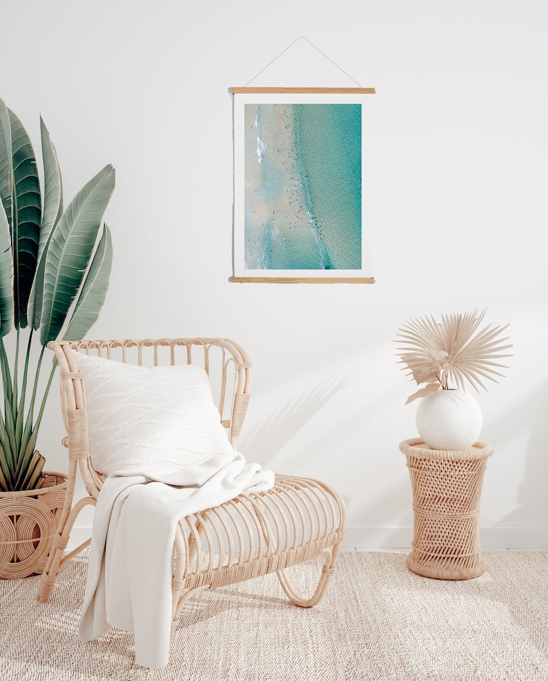 May include: A white wicker chair with a white throw blanket and a white pillow. The chair is in front of a white wall with a framed photograph of an aerial view of a beach and ocean. A small wicker side table with a white vase and a dried palm frond is to the right of the chair.