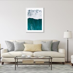 May include: A framed art print featuring an aerial view of ocean waves. The top half shows white foamy waves, transitioning to deep teal water below. The artwork is displayed above a cream-colored sofa with decorative pillows.