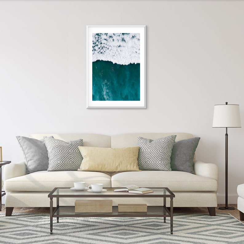 May include: A framed art print featuring an aerial view of ocean waves. The top half shows white foamy waves, transitioning to deep teal water below. The artwork is displayed above a cream-colored sofa with decorative pillows.