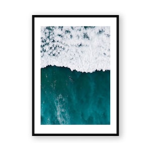 May include: Aerial view of a framed print featuring a turquoise ocean with a white foamy wave. A dark figure is visible in the water. The print is in a black frame.
