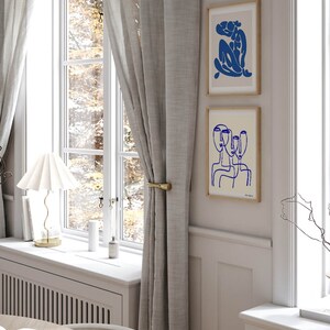 K&ouml;nnte beinhalten: Zwei gerahmte abstrakte Linienzeichnungen in Blau auf cremefarbenem Hintergrund. Die obere Zeichnung zeigt eine sitzende Figur, w&auml;hrend die untere Zeichnung drei &uuml;berlappende Gesichter zeigt.