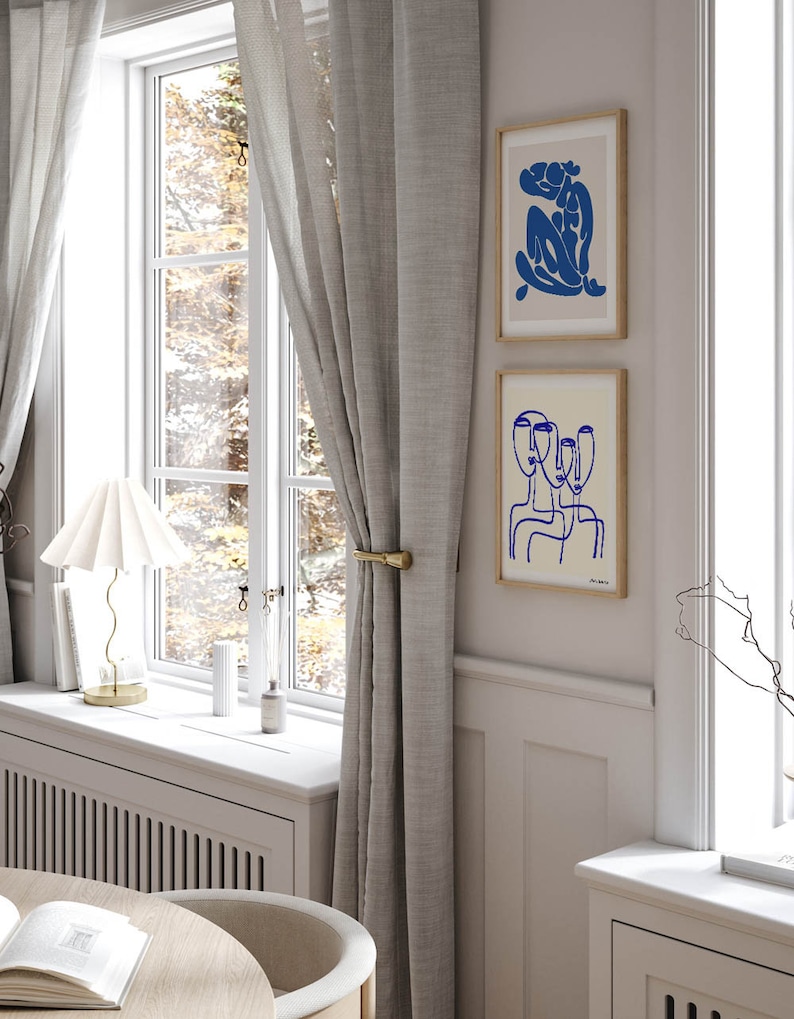 K&ouml;nnte beinhalten: Zwei gerahmte abstrakte Linienzeichnungen in Blau auf cremefarbenem Hintergrund. Die obere Zeichnung zeigt eine sitzende Figur, w&auml;hrend die untere Zeichnung drei &uuml;berlappende Gesichter zeigt.