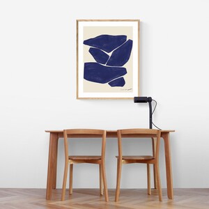 May include: A framed abstract art print featuring navy blue shapes on a cream background. The artwork is displayed above a wooden table with two matching chairs. A black desk lamp is positioned on the table.