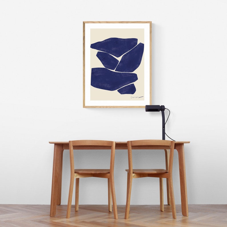 May include: A framed abstract art print featuring navy blue shapes on a cream background. The artwork is displayed above a wooden table with two matching chairs. A black desk lamp is positioned on the table.