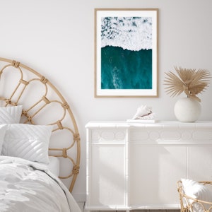 May include: A framed aerial photograph of ocean waves. The top half of the image shows white foamy waves, while the bottom half displays deep teal water. The frame is a light brown color, and the photograph is displayed on a white wall.