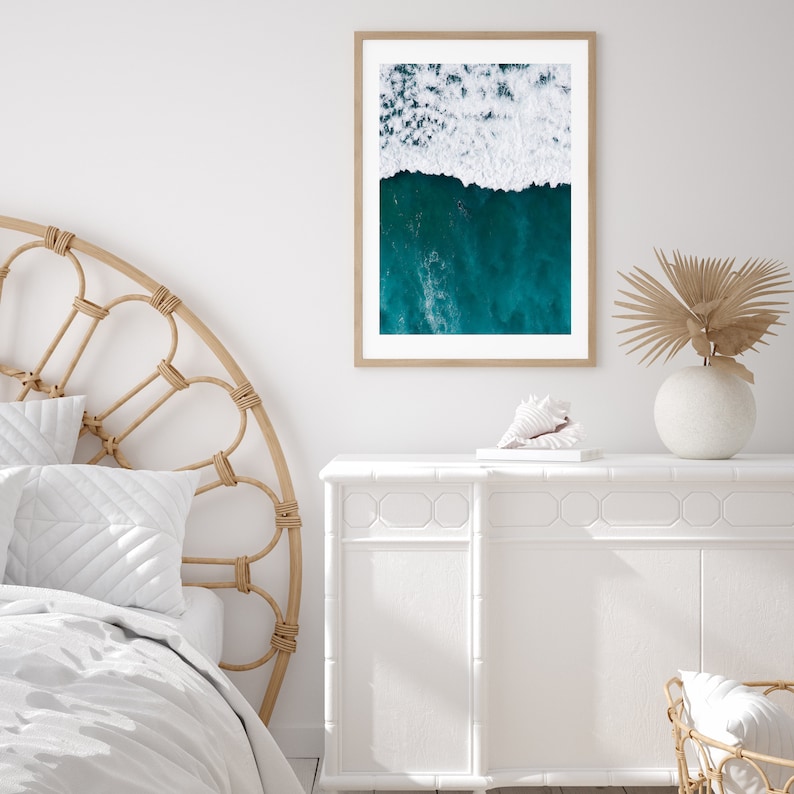 May include: A framed aerial photograph of ocean waves. The top half of the image shows white foamy waves, while the bottom half displays deep teal water. The frame is a light brown color, and the photograph is displayed on a white wall.
