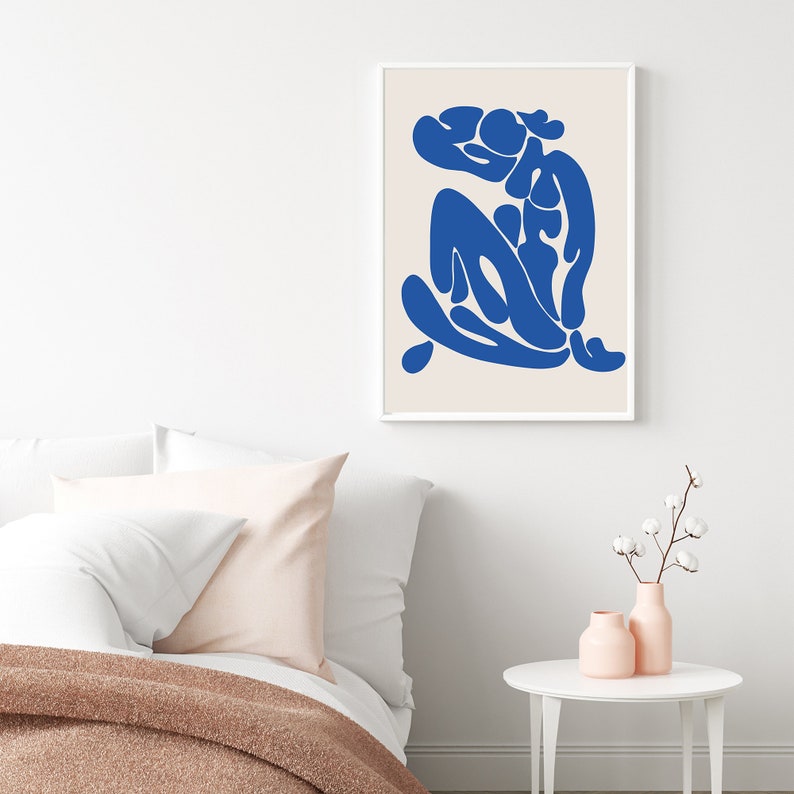 Pu&ograve; includere: Una stampa artistica astratta incorniciata con una figura blu su sfondo crema. L'opera &egrave; esposta in una cornice bianca, aggiungendo un tocco moderno. La stampa &egrave; sopra un letto con cuscini bianchi e una coperta rosa, creando un'atmosfera accogliente.