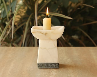 May include: A white marble candle holder with a decorative black trim. A lit candle sits in the holder.