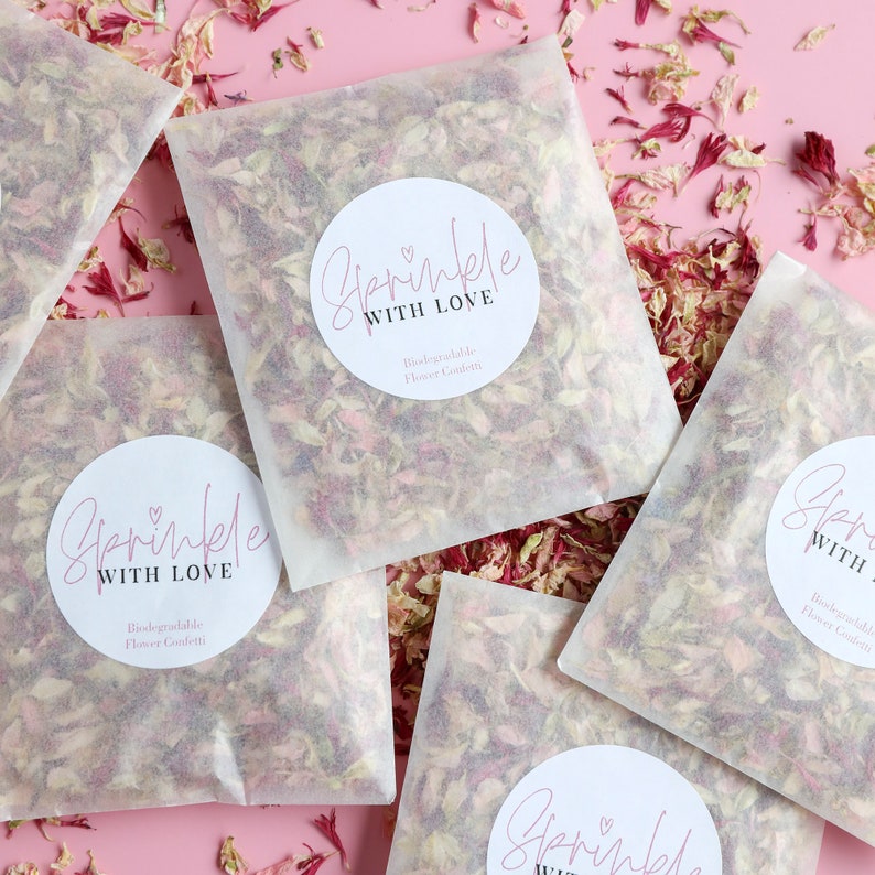 May include: Four white paper bags filled with pink and white flower petals. Each bag has a round sticker with the text "Sprinkle with Love" and "Biodegradable Flower Confetti".