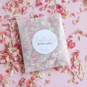 May include: A white paper bag filled with pink and white flower petals. The bag has a round sticker with the text "Sprinkle with Love" and the words "Biodegradable Flower Confetti" underneath.