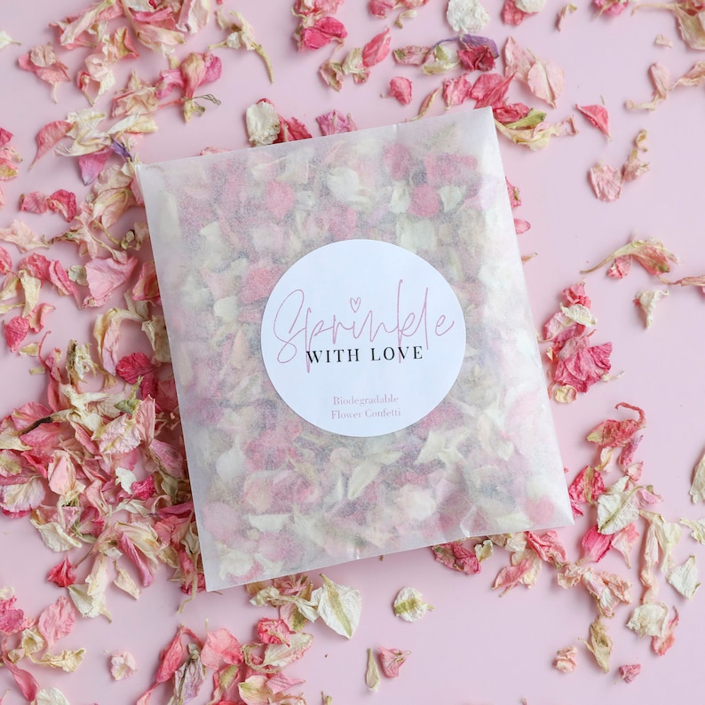 May include: A white paper bag filled with pink and white flower petals. The bag has a round sticker with the text "Sprinkle with Love" and the words "Biodegradable Flower Confetti" underneath.