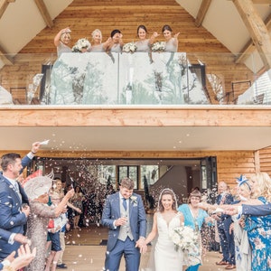 May include: A bride and groom walk down the aisle after their wedding ceremony, surrounded by guests throwing confetti. The bride is wearing a white wedding dress and the groom is wearing a dark suit. The bridesmaids are standing on a balcony above the couple, throwing confetti.